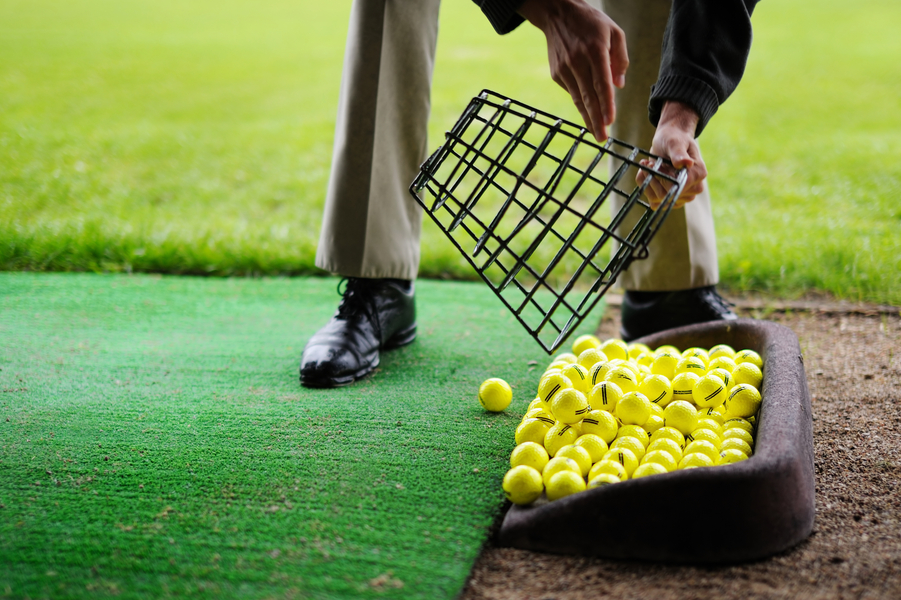 A man empties a bucket of bright yellow practice golf balls into a bin adjacent to a driving mat.
