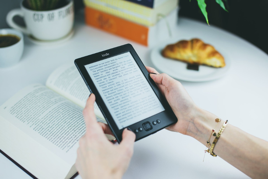 Hands holding a Kindle device in front of an open book, with coffee, and a croissant